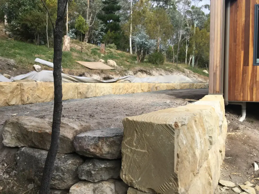 Sandstone boulder retaining wall corner beside the house, with landscape fabric laid on the batter above