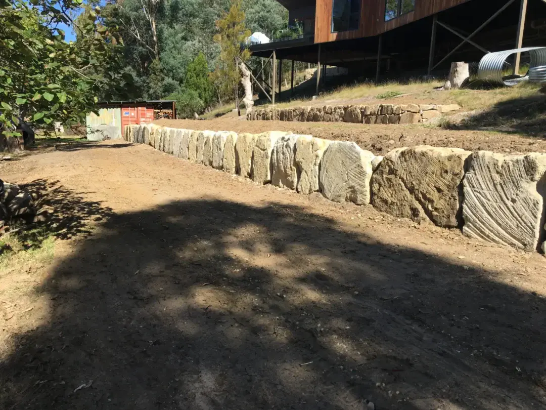 Two-tier sandstone boulder retaining wall under construction beside the new driveway