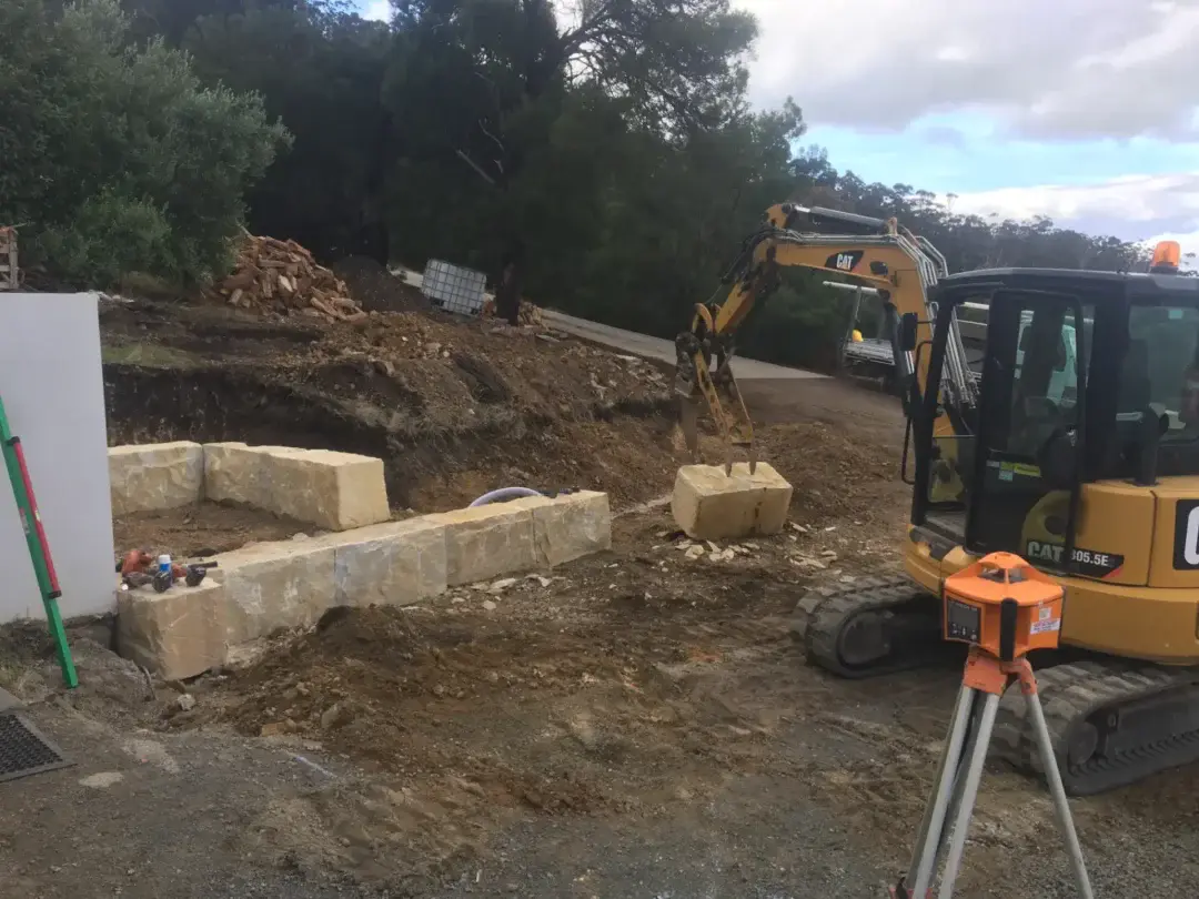Excavator placing sandstone blocks for a new retaining wall beside a rendered boundary wall