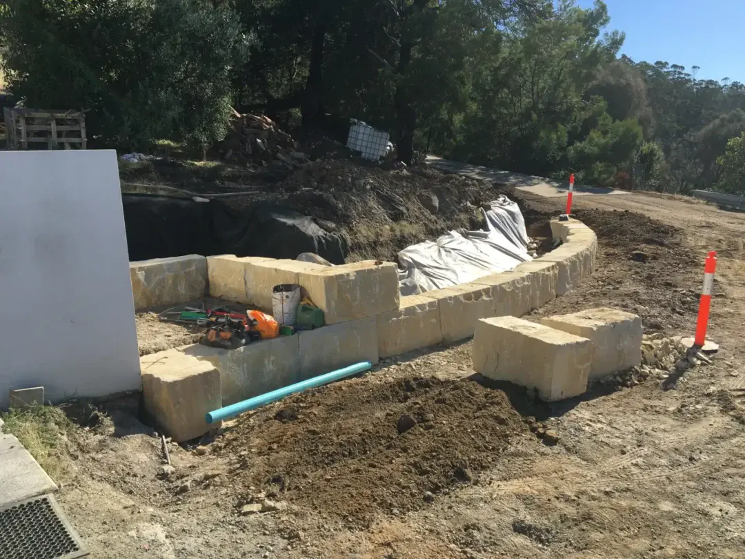Sandstone block retaining wall under construction beside a roadside garden bed, stormwater pipe in place