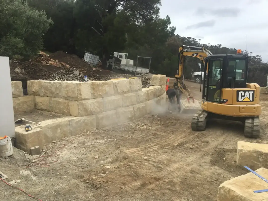 Two-tier sandstone block retaining wall being built, excavator and worker on site